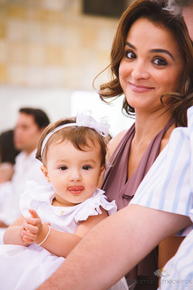 Criança mostrando a língua, sentada no colo da mãe dentro da igreja, durante seu batizado.