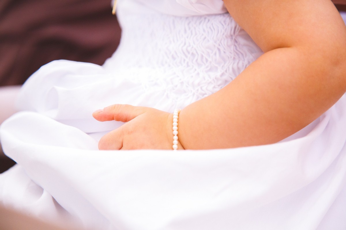 Detalhe da mão e pulseira da criança, sentada no colo do pai durante seu batizado.