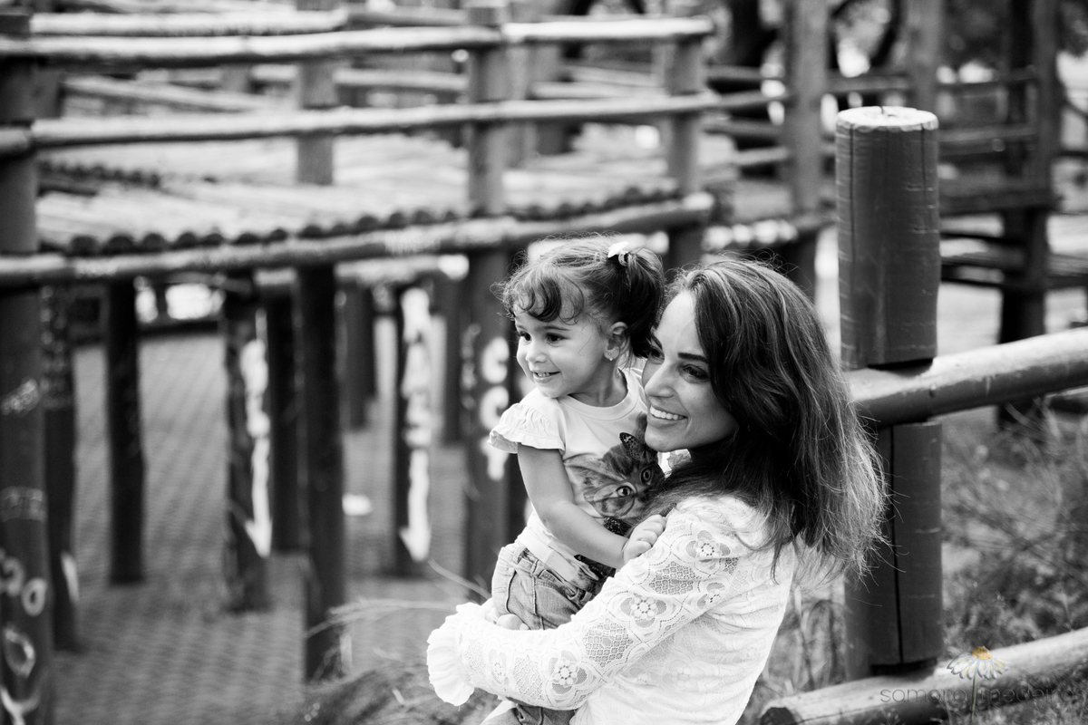 Foto em preto e branco de uma criança no colo de sua mãe distraídas com a paisagem em uma praça.