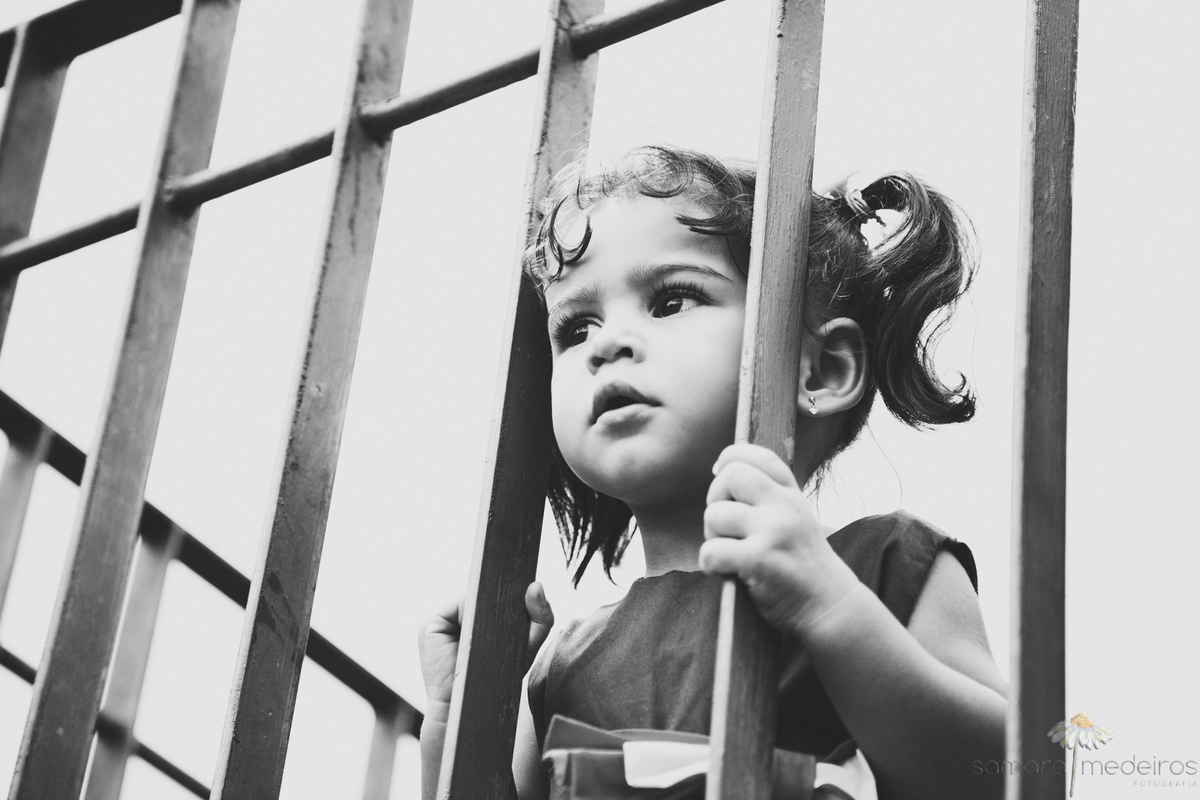 Foto em preto e branco de uma criança observando as pessoas brincando em um parque entre as grades da cerca do mirante.