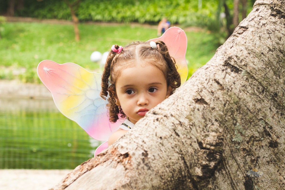 Criança se escondendo atrás de uma árvore em um ensaio externo no parque Municipal de Belo Horizonte.