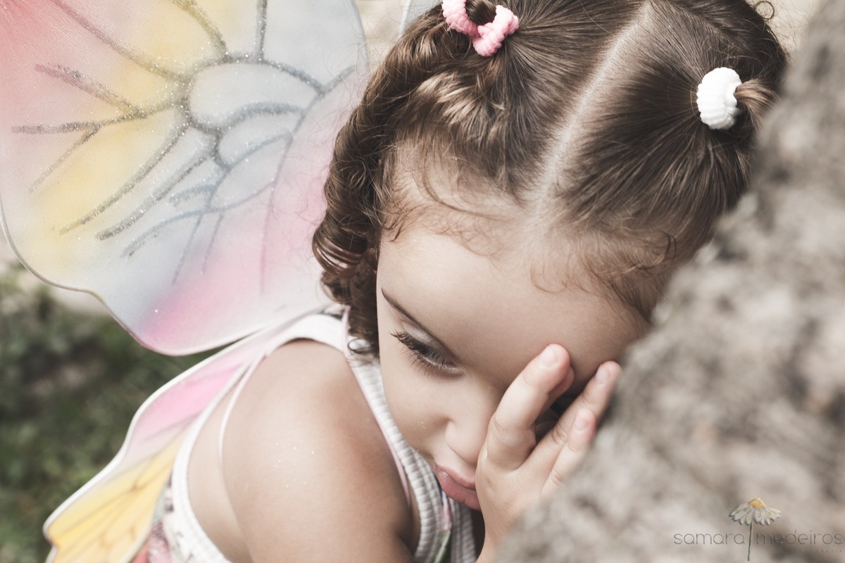 Criança com asas de fada apoiada em uma árvore, durante uma sessão fotográfica em Belo Horizonte.