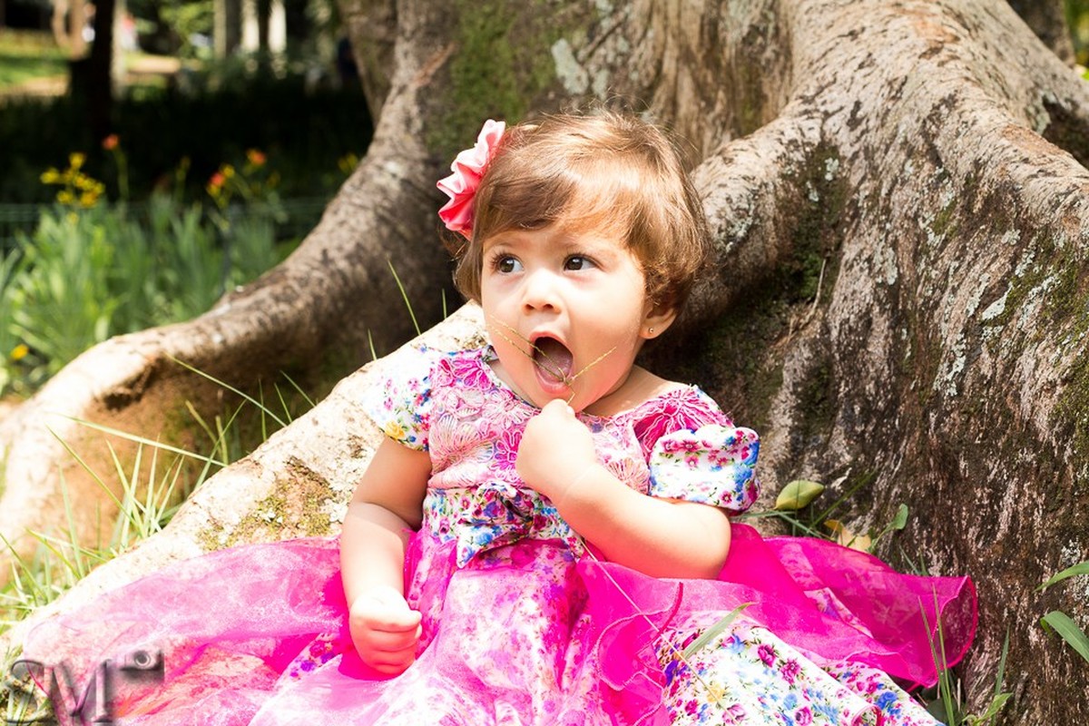 Criança em um ensaio externo em Belo Horizonte, apoiada em uma árvore tentando comer um capim