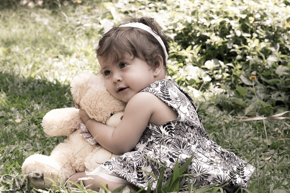 Criança sentada na grama abraçando um ursinho de pelúcia, em ensaio externo em um parque de Belo Horizonte.