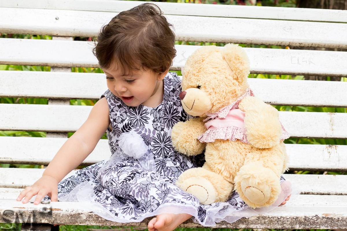 Criança com ursinho de pelúcia abraçada, sentada em um banco de praça, em ensaio infantil externo feito em um parque de Belo Horizonte, BH.