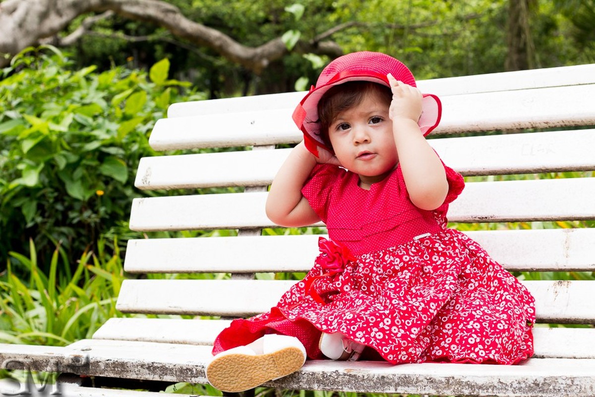 Criança sentada em um banco de parque segurando um chapéu vermelho em sua cabeça, em ensaio externo infantil feito em um parque em Belo Horizonte, BH.