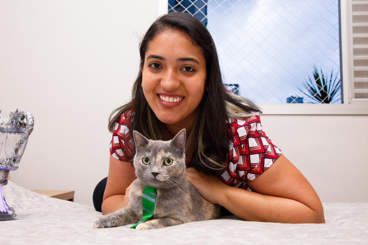 Gata cinza com uma gravata verde da Sonserina deitada na cama com sua totura atrás dela dando um abraço, ambas olhando para a foto.