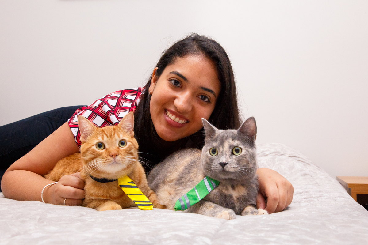 Tutora deitada na cama com seus dois gatos, um amarelo e uma cinza, dando um abraço em ambos.
