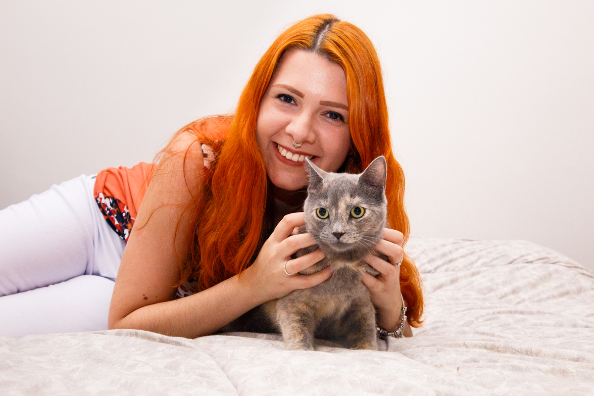 Tutora deitada na cama, olhando para a câmera e sorrindo e abraçando sua gatinha cinza.