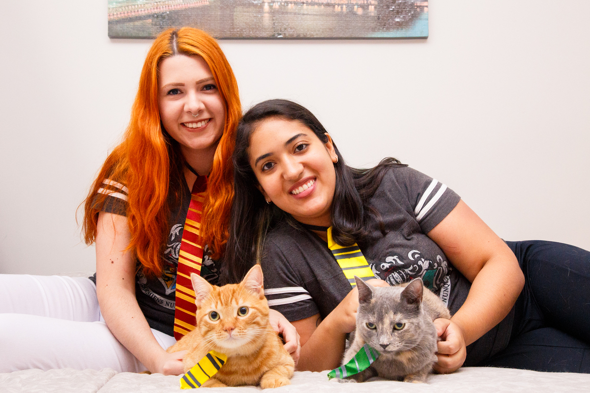 Casal sentadas na cama com os dois gatinhos em frente a elas, todas usando gravatas de Hogwarts.