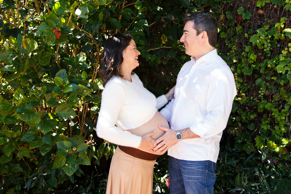 Gestante de pé sorrindo para seu marido que está de frente para ela, ambos se olhando, ao fundo folhagens de árvores.