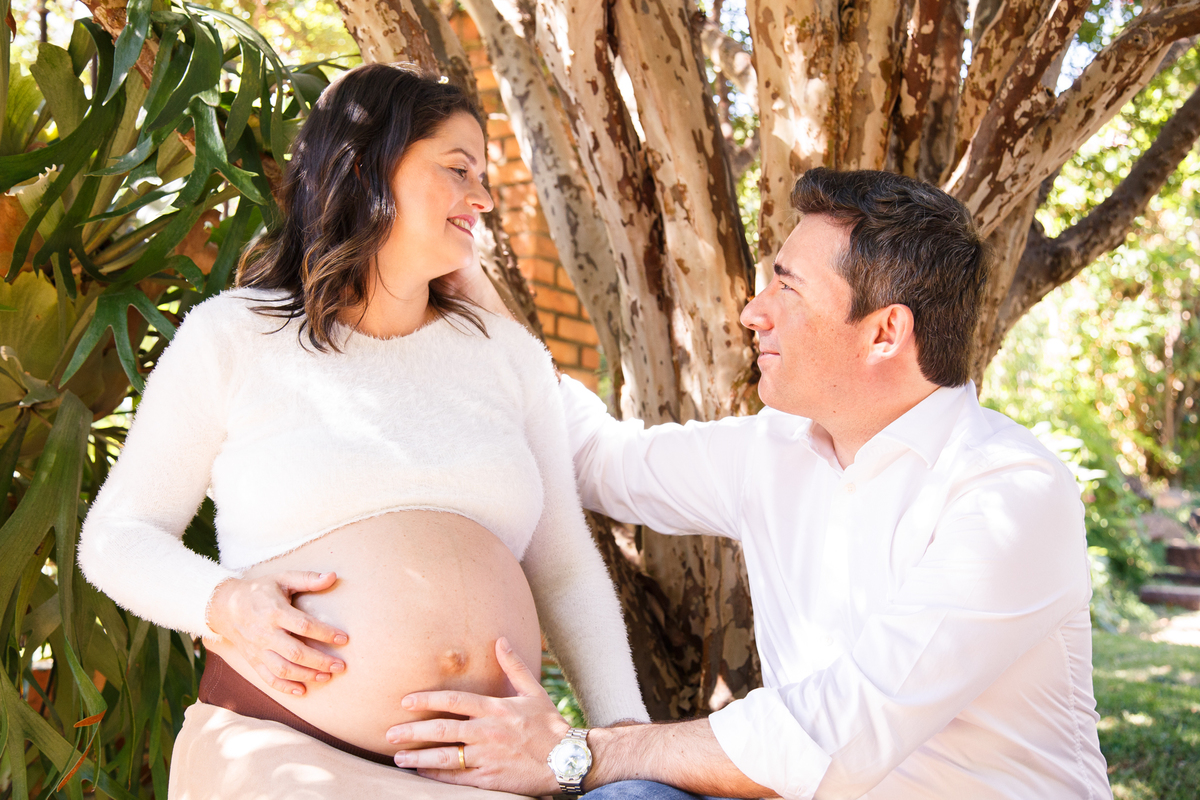 Casal sentado embaixo de uma jabuticabeira, olhando um para o outro e sorrindo, ambos com a mão na barriga da mulher que está grávida.