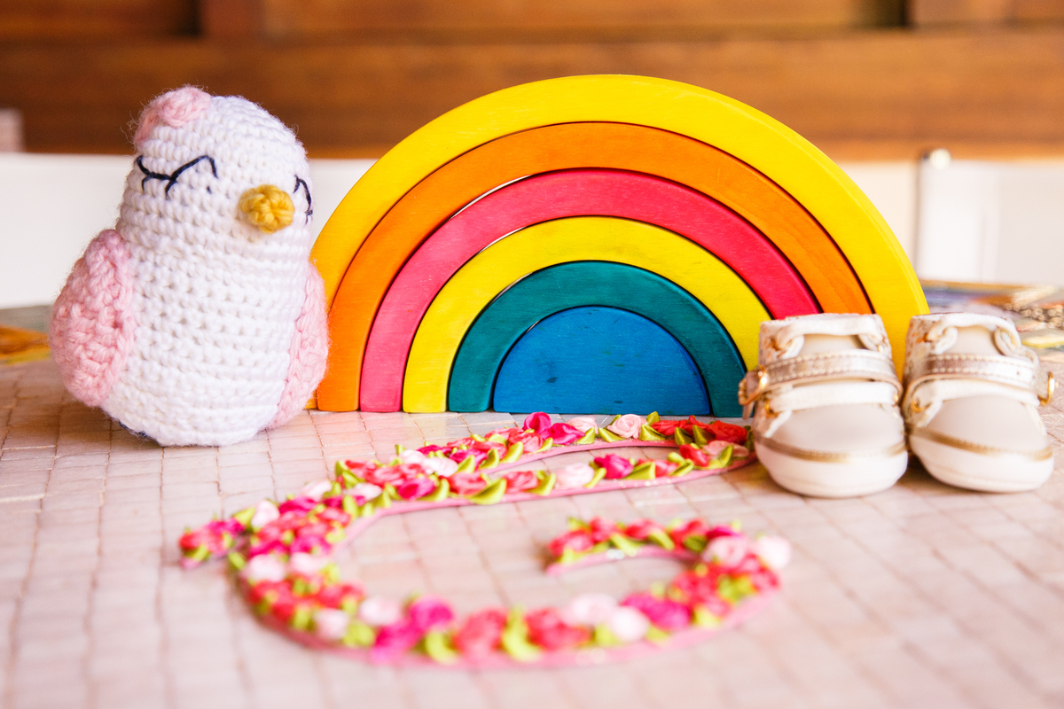 Decoração do quarto da bebê que está para chegar, com um arco-íris, um passarinho tipo amigurumi e um C coberto de florzinhas. E um sapatinho da bebê ao lado.