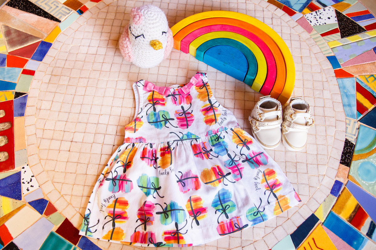 Foto de cima da decoração do quarto da bebê sobre uma mesa de mosaicos, um arco íris, um passarinho no estilo amigurumi, um sapatinho e um vestido branco com coloridinhos.