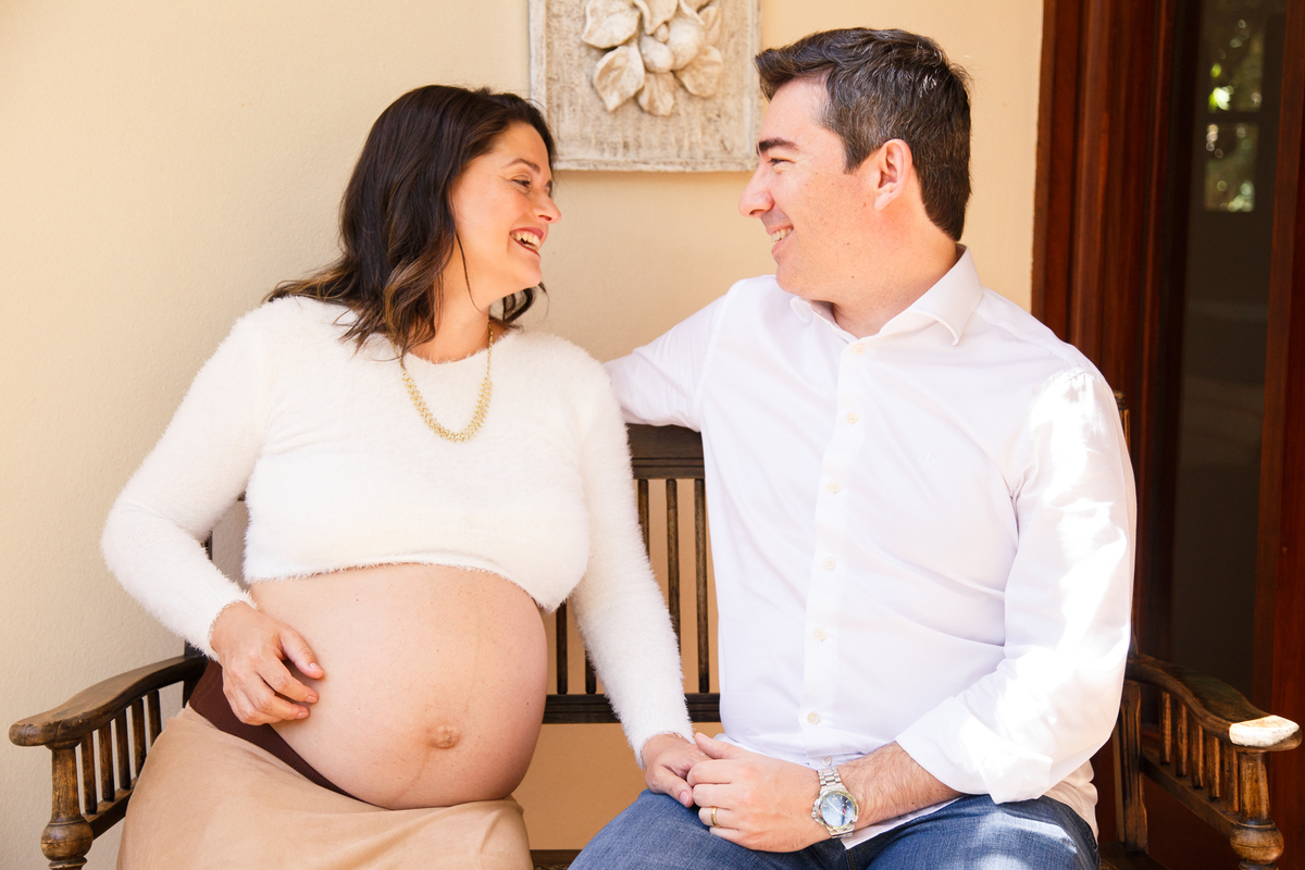 Casal sentado em um banco em sua casa, se olhando e sorrindo, a moça com a mão na barriga esperando sua bebê.