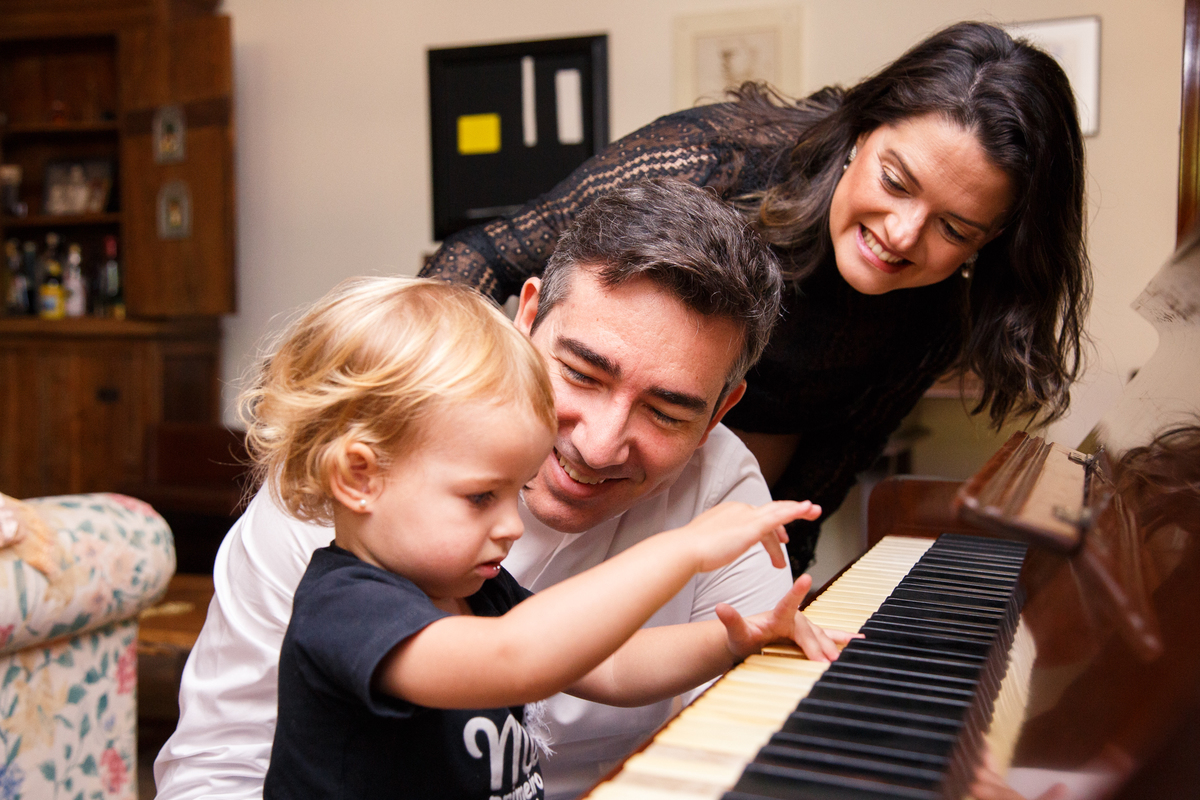 Foto da família com uma criança sentada ao piano brincando de tocar e os pais agachados ao seu lado observando e sorrindo.