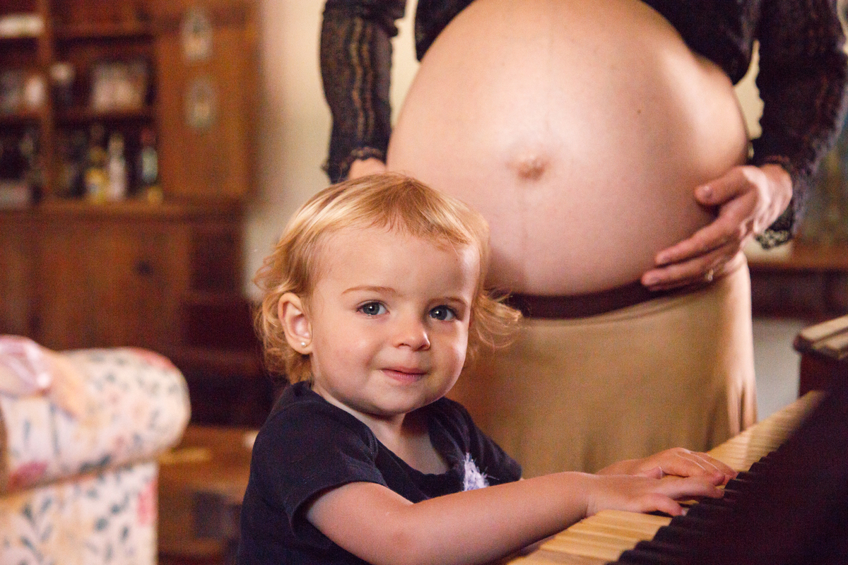 Criança sorrindo, olhando para a câmera, brincando que está tocando piano com a barriga grávida de sua mãe atrás.