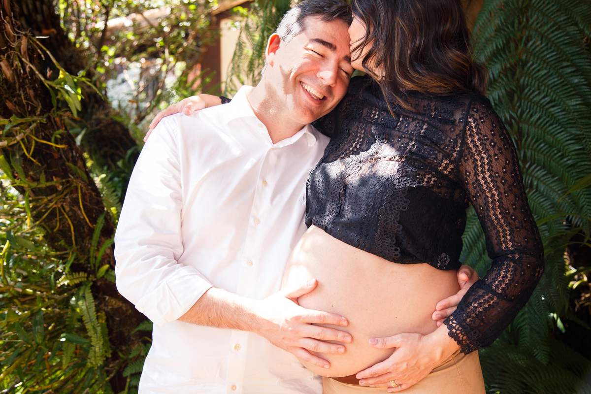 Casal abraçado, o homem deitado no ombro da mulher sorrindo enquanto ela dá um beijo em sua testa, ambos com uma mão apoiada na barriga dela, gestante.