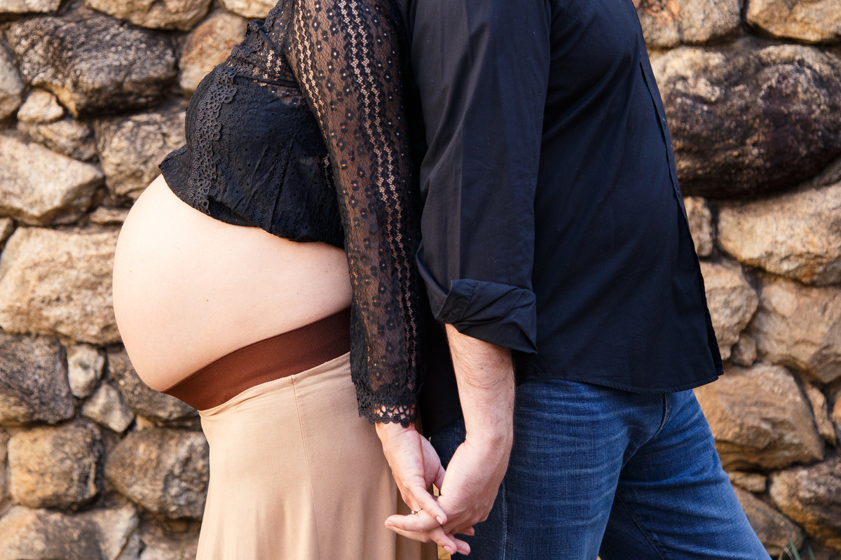 Foto do ombro para baixo de um casal com ênfase na barriga da moça, que está grávida. Eles de costas um para o outro, com os dedos entrelaçados.