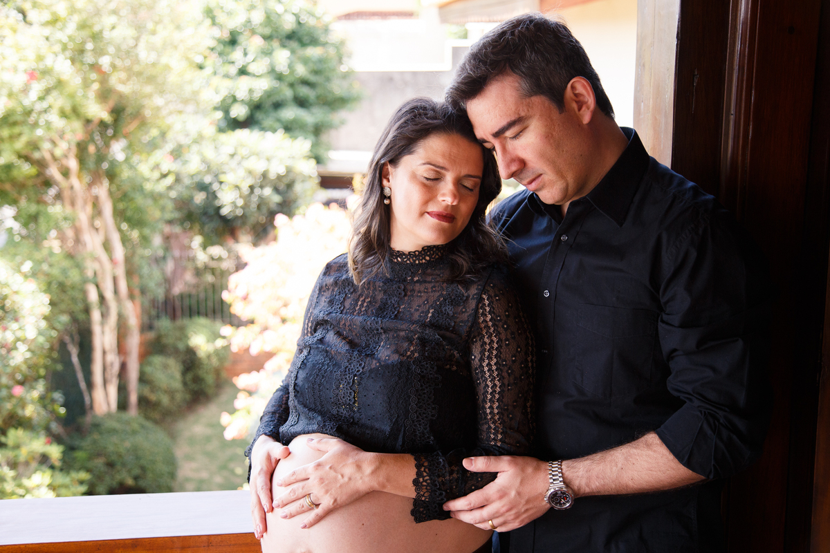 Casal encostado um no outro, a moça que está grávida com as costas apoiada no peito do marido, ambos de olhos fechados com as cabeças se tocando.