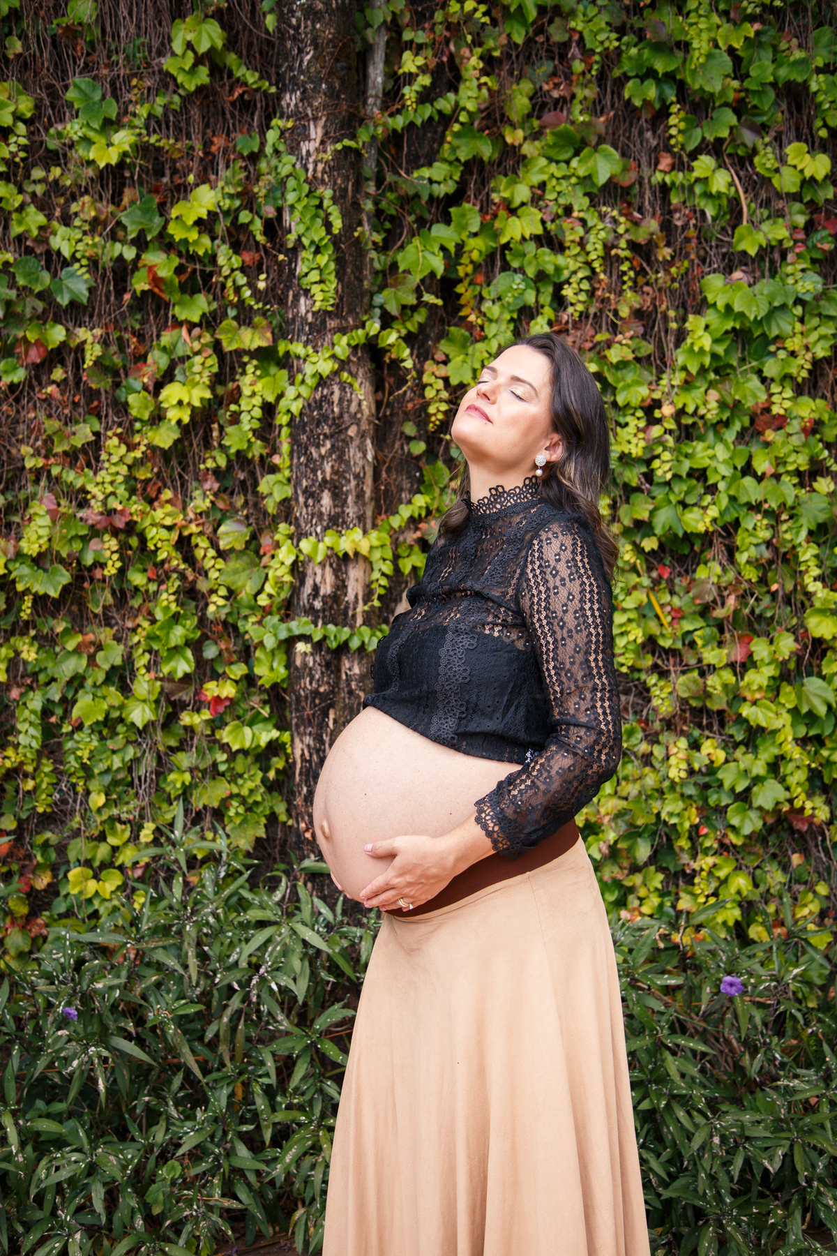 Gestante com a mão apoiada na barriga, a cabeça virada para cima, com os olhos fechados e uma expressão pensativa. Atrás dela um muro coberto de trepadeiras.