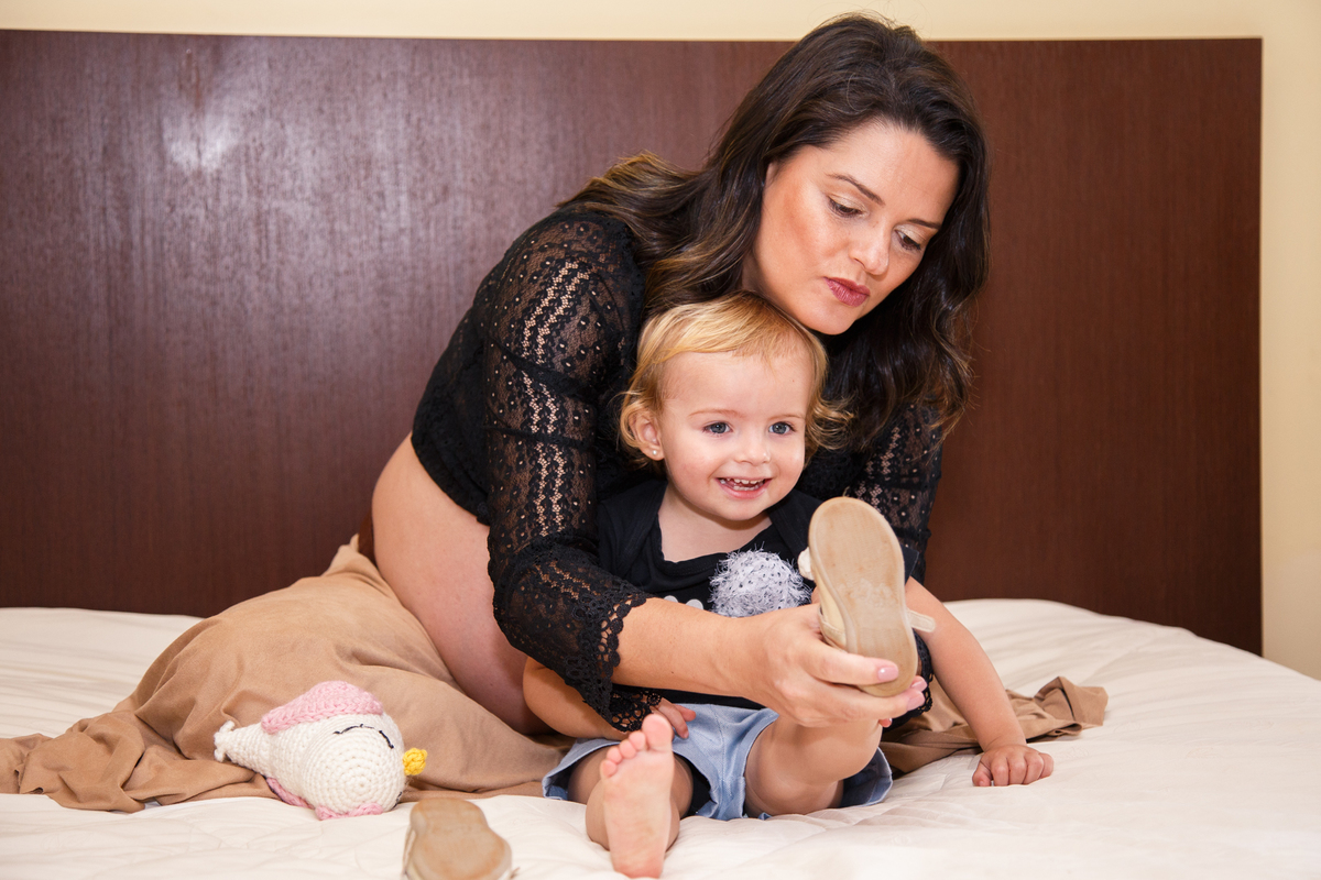 Mãe grávida do segundo bebê tirando o sapatinho da primeira filha em cima de uma cama.