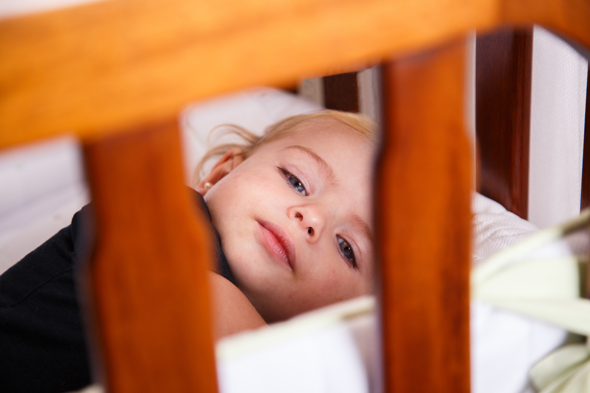 Criança deitada em um berço, olhando para a câmera por entre as barras de madeira da grade do berço.