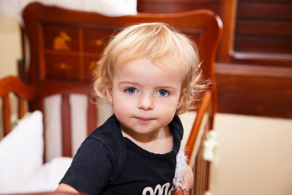 Criança dentro de um berço de madeira olhando para a câmera com uma expressão séria.