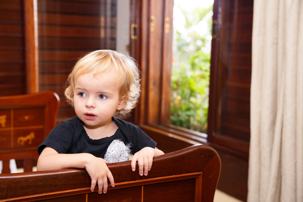 Criança em pé dentro de um berço olhando para a mãe que está ao lado e não aparece na foto, com janelas de madeira ao fundo, mostrando parte do jardim lá fora.