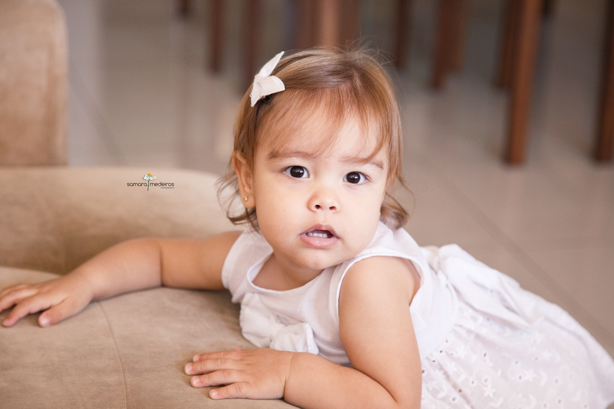 Criança de roupinha branca, lacinho no cabelo apoiada no sofá de sua casa durante ensaio fotográfico de família.