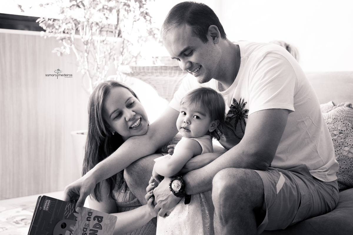 Foto em preto e branco da família reunida para ler um livro, a bebê nos braços do pai e a mãe sentada ao lado deles. A bebê olha para trás por sobre o ombro para a foto.