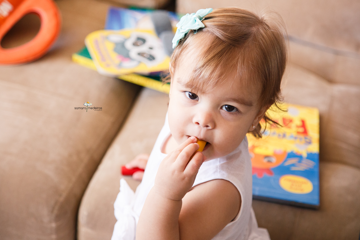 Foto de um ângulo de cima de uma bebê apoiada no sofá de sua casa colocando um brinquedinho na boca.
