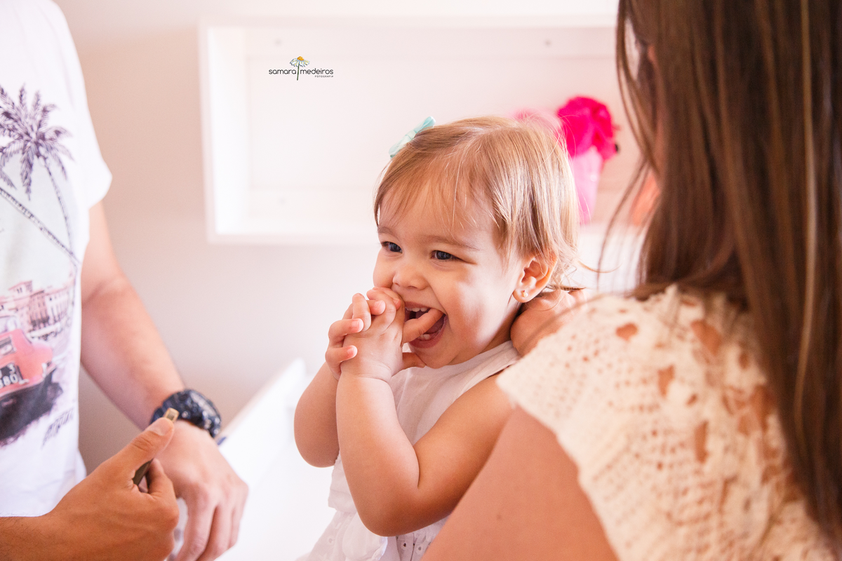 Bebê sentada no trocador de seu quarto com as mãozinhas na boca coçando os dentes que estão nascendo.