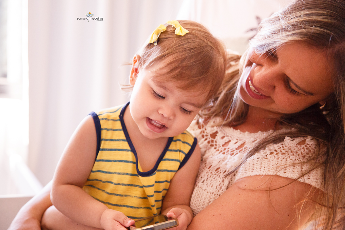 Mãe abraçando sua bebê e olhando para ela, enquanto a bebê sorri olhando para um celular em sua mão.