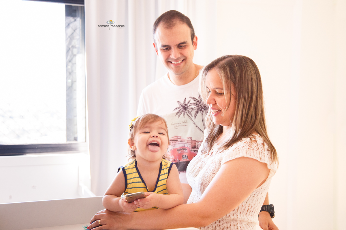 Mãe, pai e bebê em seu quartinho, trocando de roupa, pai e mãe olhando para a bebê enquanto ela olha para a avó, que não aparece na foto e faz uma careta rindo.