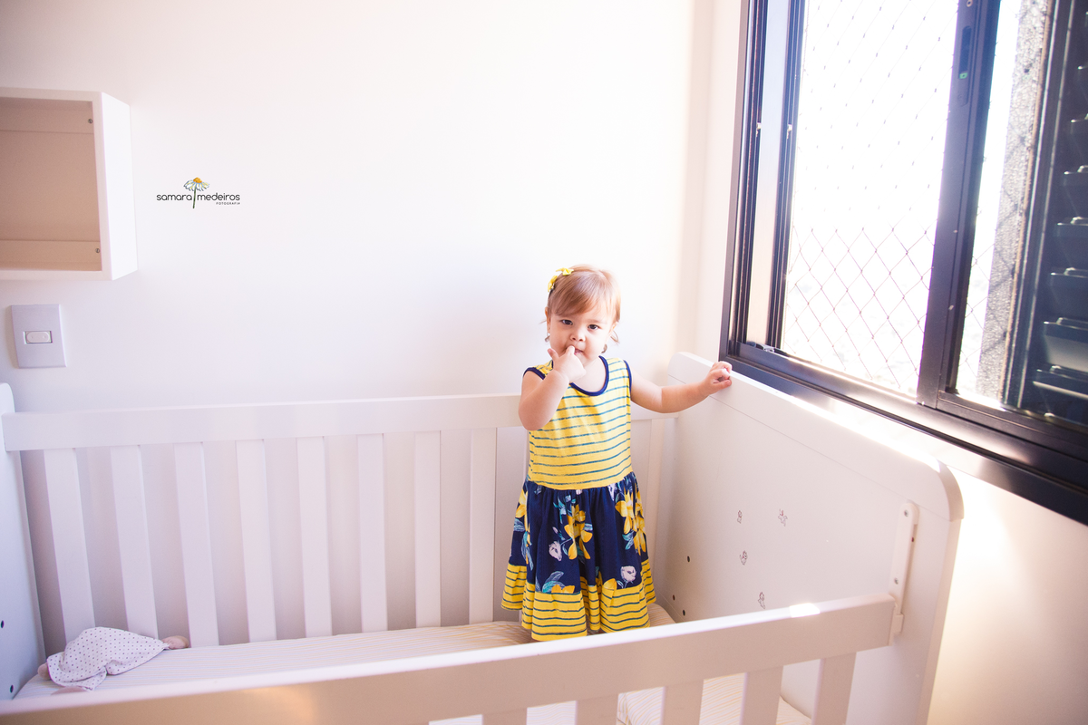 Foto aberta de uma bebê em seu berço, ela está em pé com um vestidinho amarelo e azul com a mão apoiada na janela e a outra mão na boca coçando os dentes.