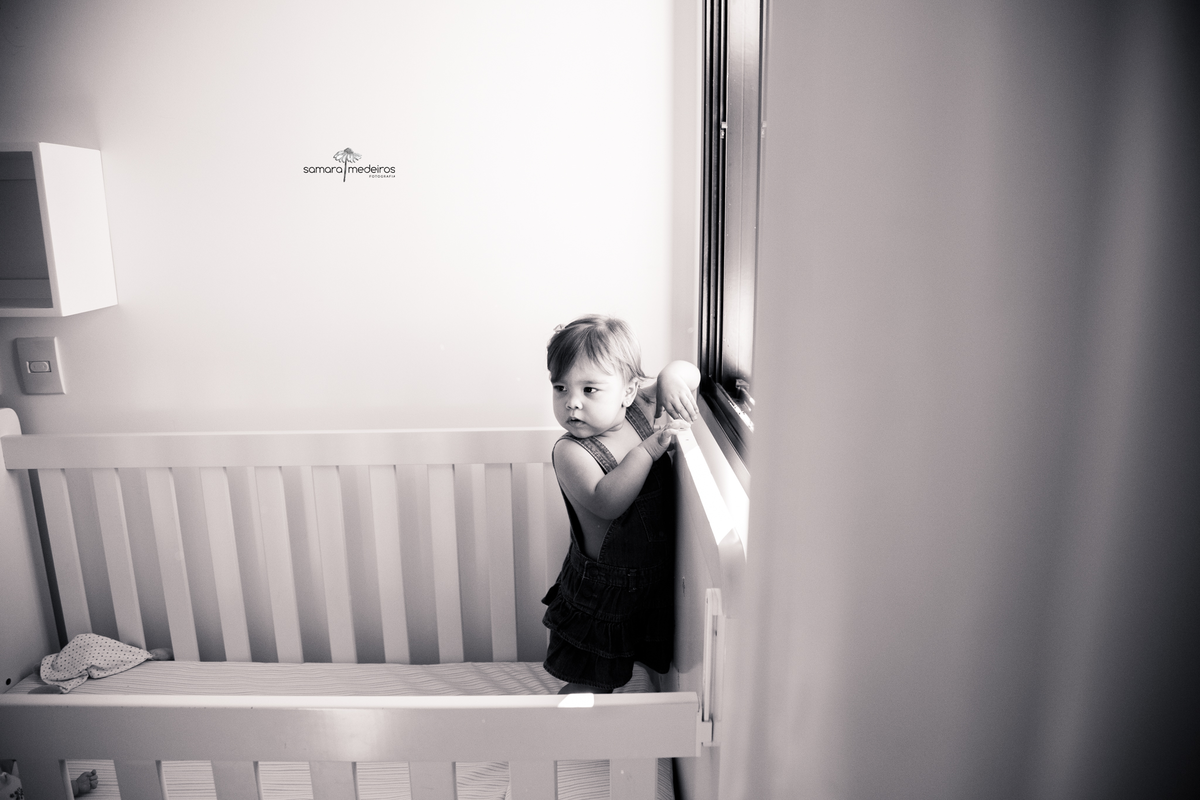 Foto em preto e branca de uma criança de pé em seu berço, com as duas mãos na janela para se apoiar e o olhar direcionado para trás para ver os pais.
