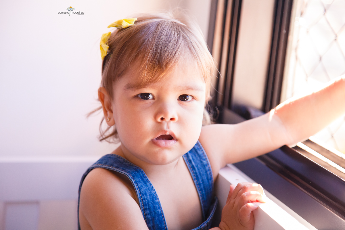 Close no rosto de uma bebê que está apoiada em uma janela que fica aos pés de seu berço. Ela olha para a câmera.