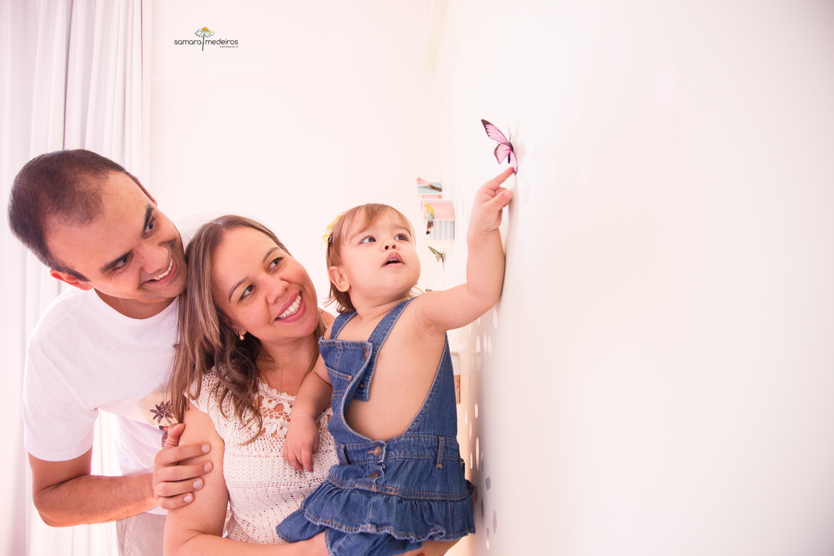 Pai e mãe com sua bebê no colo olhando para uma borboleta de enfeite na parede de seu quarto.