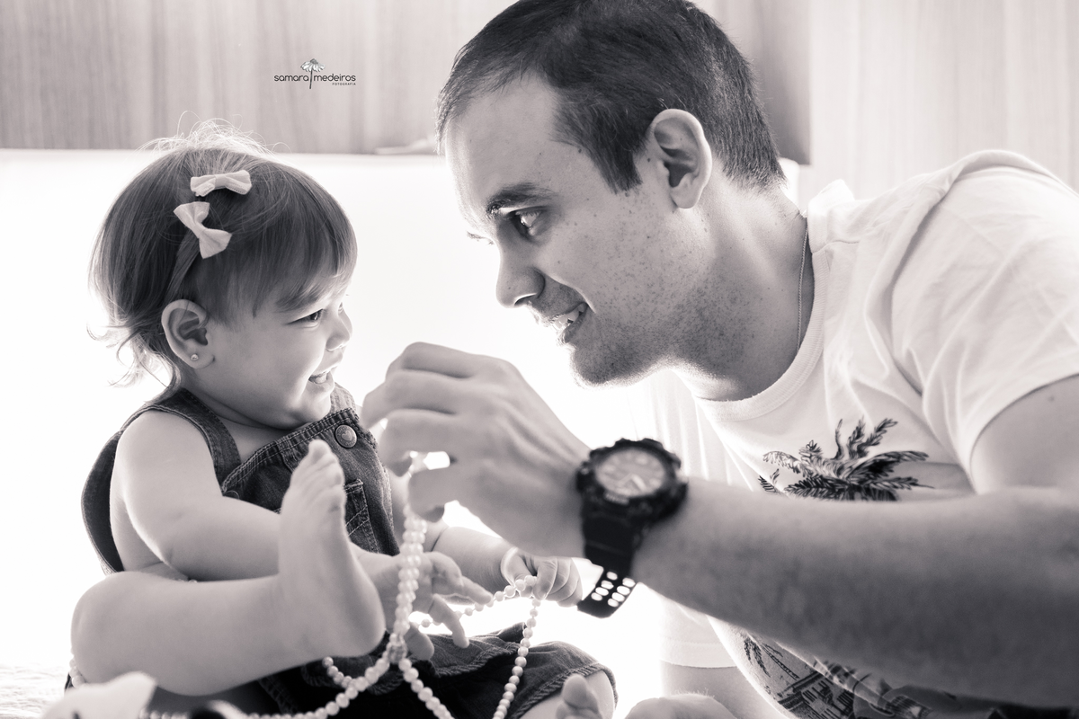 Foto em preto e branca de uma criança brincando com seu pai na cama, a bebê brinca com um colar de pérolas da mãe, eles se olham e sorriem.