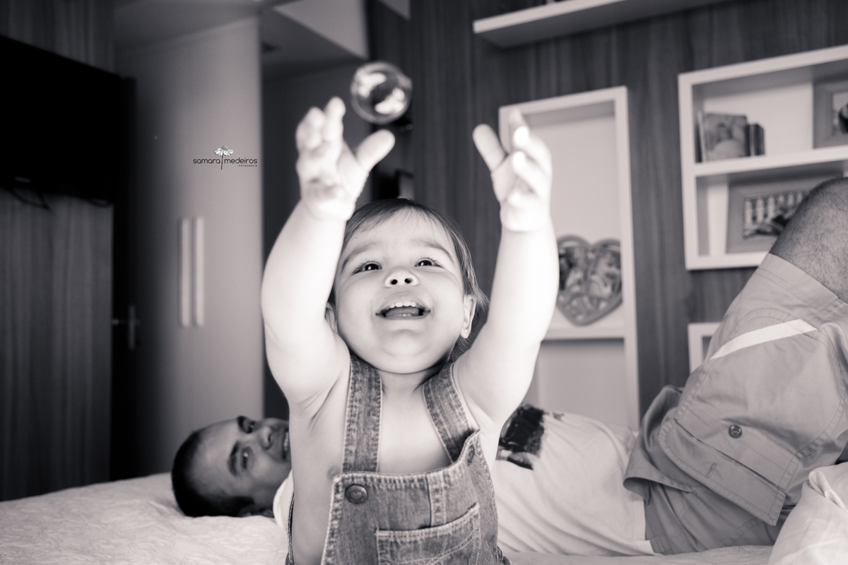 Foto em preto e branco de uma bebê brincando de pegar bolinhas de sabão, com um sorriso. O pai a observa ao fundo da foto.
