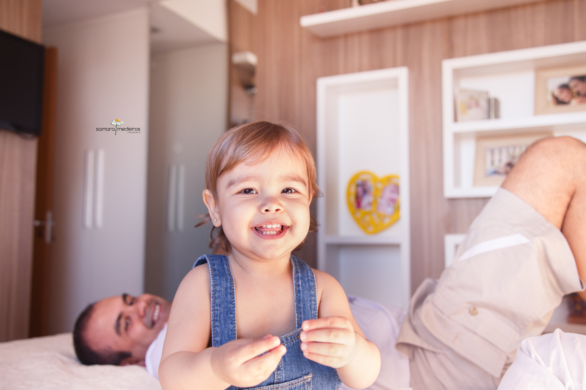 Bebê sentada na cama dos seus pais rindo e brincando com a mãe com o pai ao fundo deitado, observado ela e sorrindo.