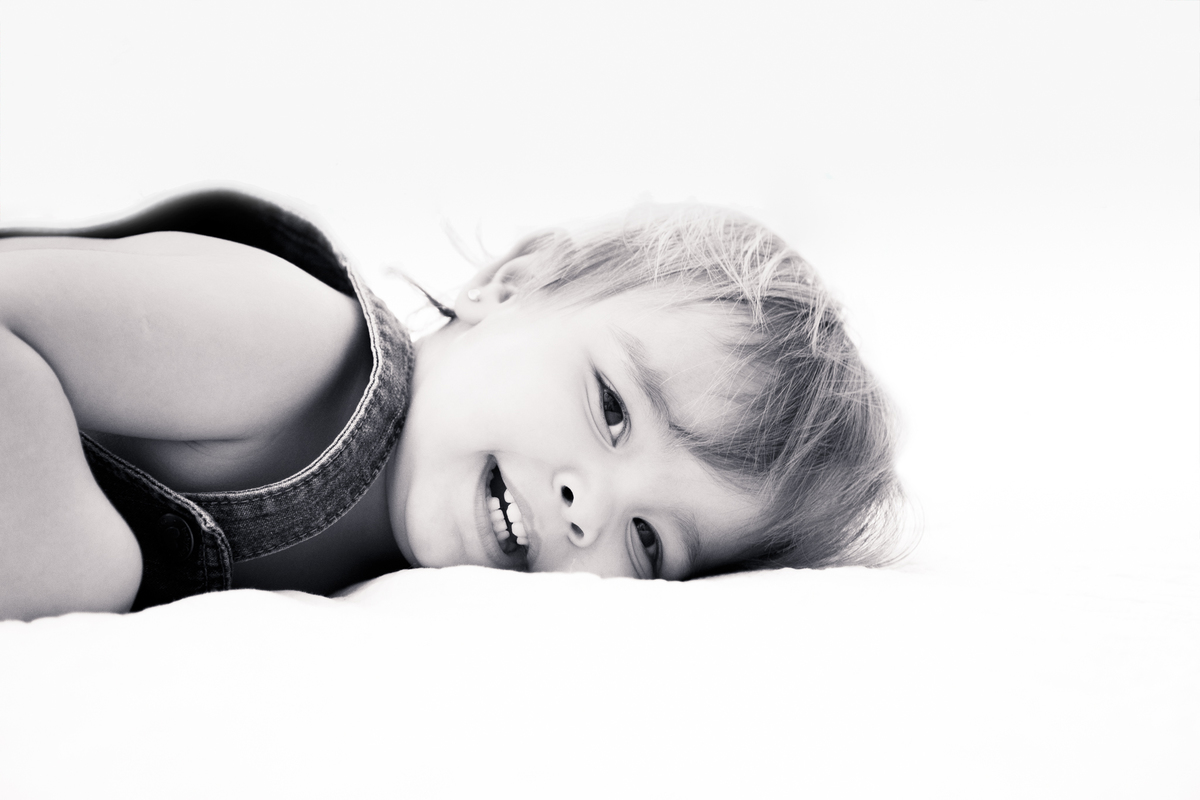 Foto em preto e branco de uma bebê deitada de lado na cama dos pais sorrindo e olhando para a câmera.