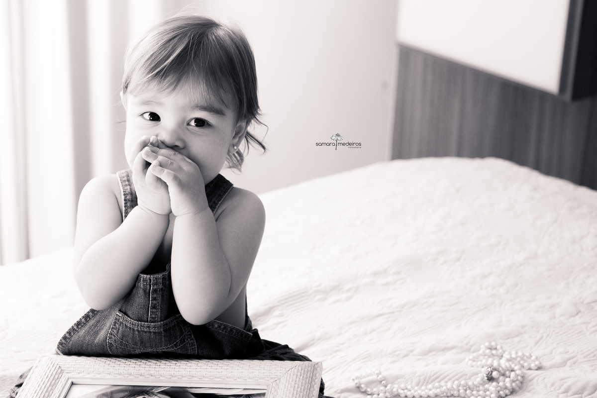 Foto em preto e branco de uma bebê sentada na cama dos pais com uma foto no colo e as mãos cobrindo a boca.