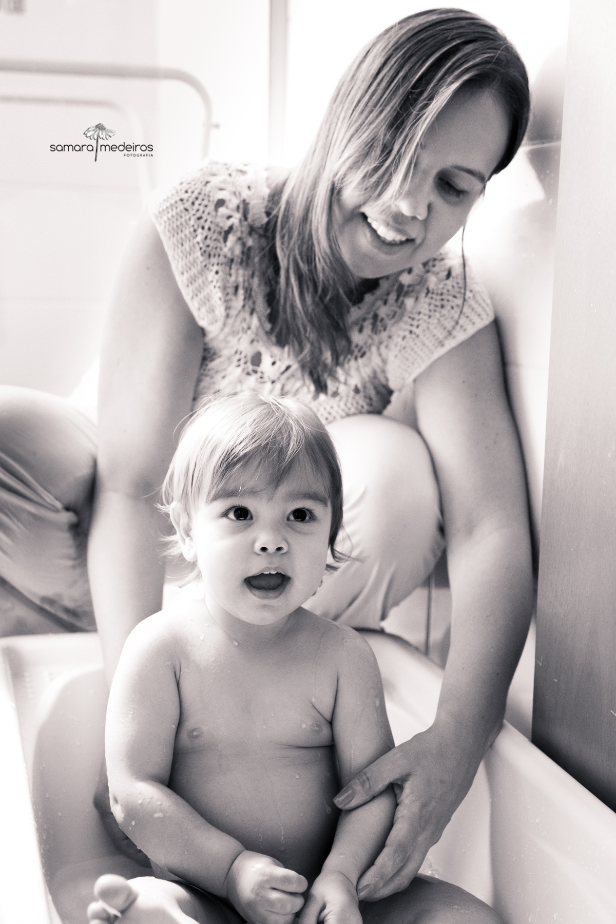 Foto em preto e branco de uma mãe dando banho em sua bebê dentro da banheirinha dela. A bebê tem uma expressão alegre e a mãe sorri.
