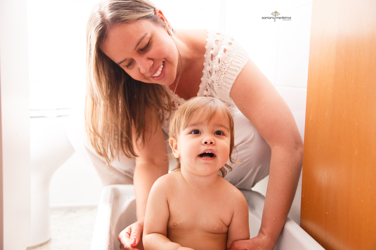 Bebê dentro de sua banheirinha tomando banho, dado por sua mãe. A mãe olha para a bebê e sorri, enquanto ela olha para a avó que não aparece na foto.
