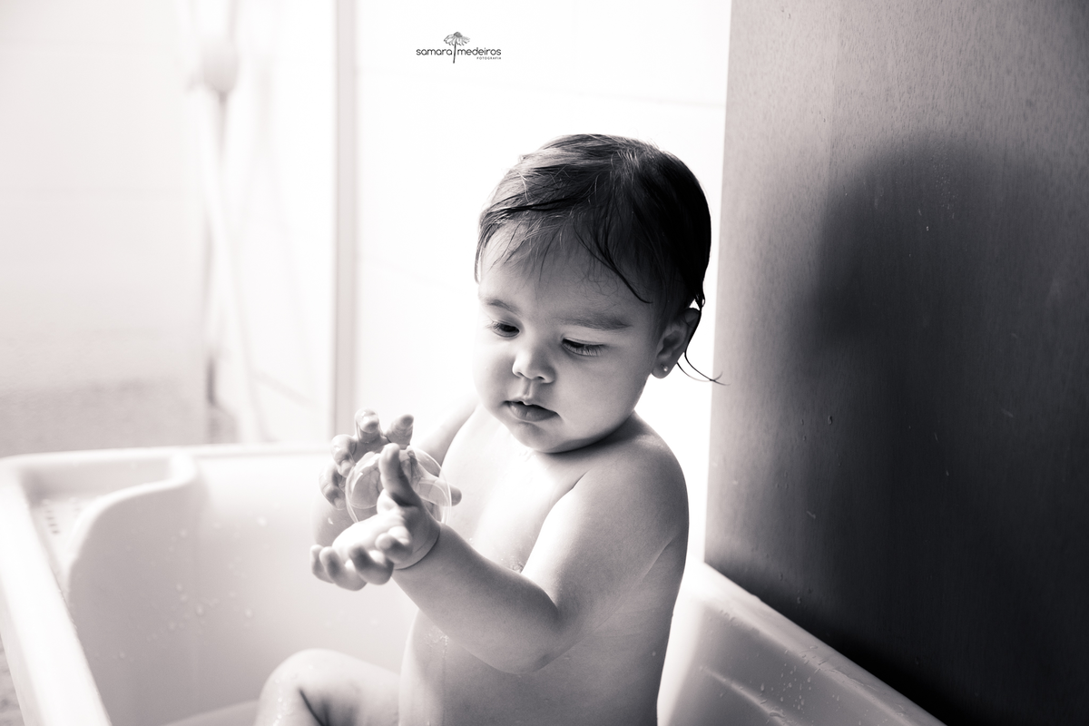 Foto em preto e branco de uma bebê dentro de sua banheirinha brincando com bolhas de sabão.