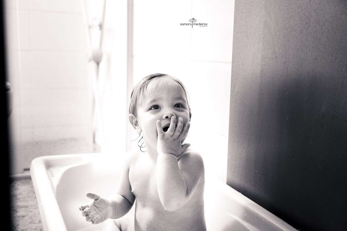 Foto em preto e branca de uma bebê em sua banheirinha tomando banho, com a mão sobre a boca, pois a avó, que não aparece na foto, está fazendo bolhas de sabão. 