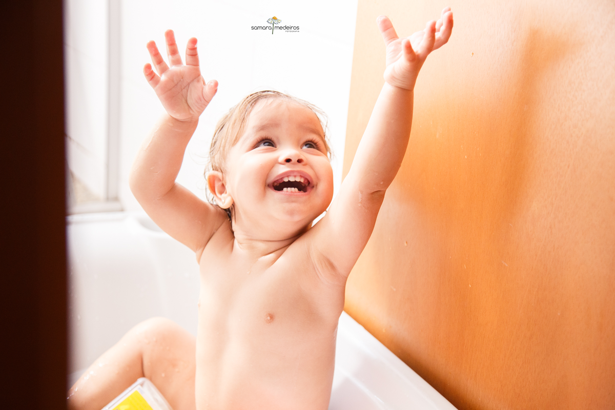 Bebê dentro de sua banheirinha com os bracinhos esticados tentando pegar uma bolha de sabão e com um sorriso enorme.