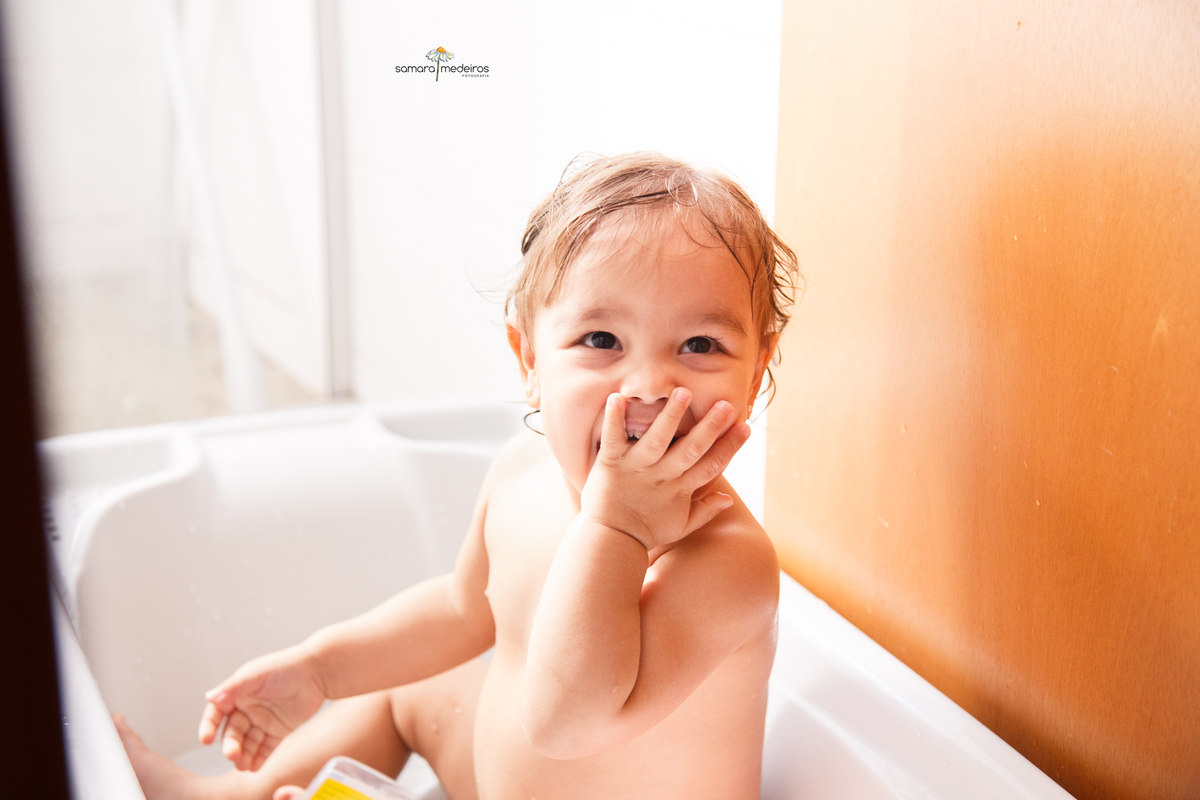 Bebê em sua banheirinha com uma mão cobrindo a boca, dando uma gargalhada, enquanto olha sua avó, que não aparece na foto, fazendo bolhinhas de sabão.